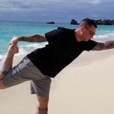 A man stretches on a beach with turquoise water and cloudy sky.