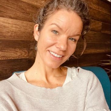 Smiling woman with curly hair in a cozy sweater against a wooden wall.