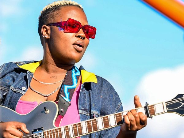 Person playing electric guitar outdoors wearing red sunglasses and denim jacket.