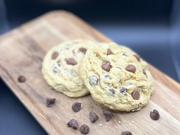 Two chocolate chip cookies on a wooden board with scattered chocolate chips.