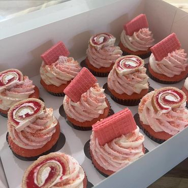 Box of pink and white swirled cupcakes topped with wafers and cake rolls.