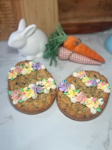 Two decorated Easter-themed cookie cakes with icing flowers, a bunny figurine, and fabric carrots in the background.
