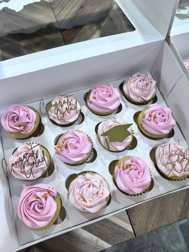 Box of pink rose-themed graduation cupcakes with congratulatory toppers.