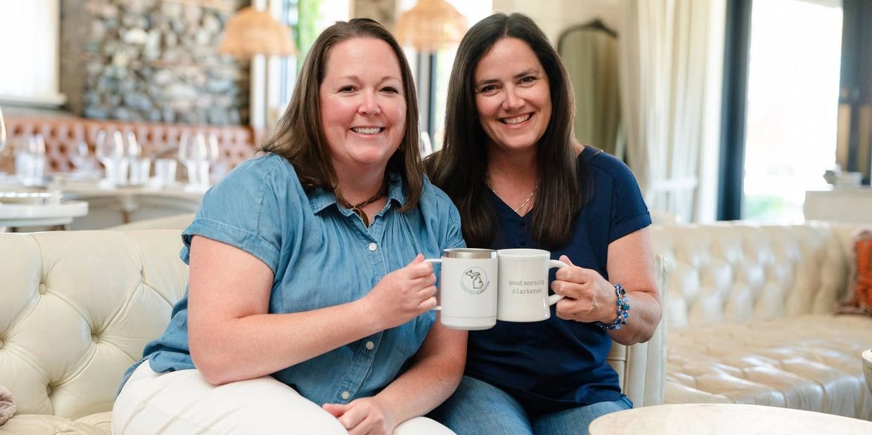 Two women smiling and toasting coffee mugs on a cozy couch.