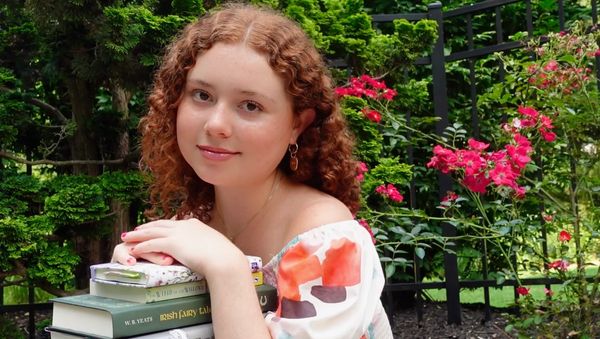 A young woman with curly red hair poses with books in a garden full of green plants and pink flowers.