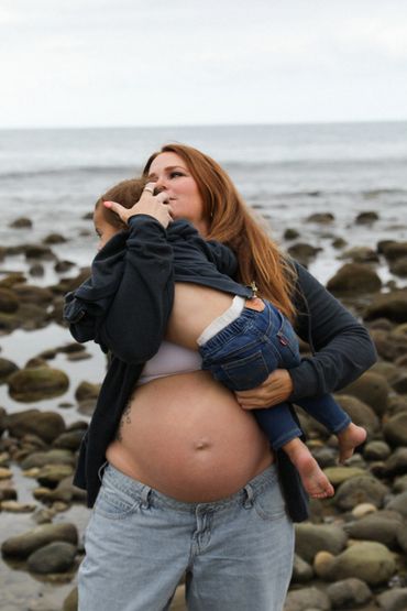 Pregnant woman holding child near rocky shore, sharing a tender moment.