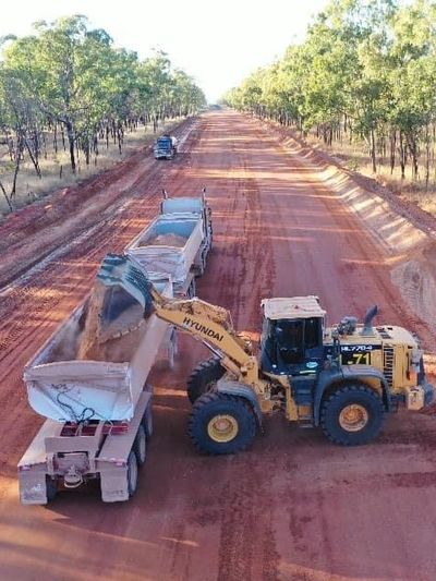 A Hyundai loader fills a dump truck on a red dirt road through a forest.