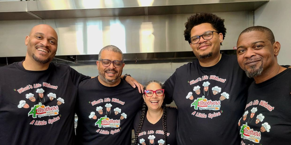 Five people smiling in matching '4 Brothers and A Lady' spice company shirts in a kitchen.