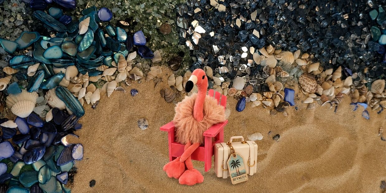 Plush flamingo on a pink chair with a suitcase on a sandy beach.