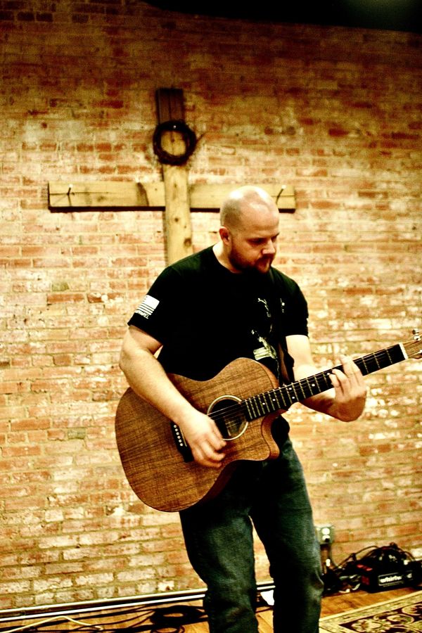 A guy on stage in a church in front of a cross playing acoustic guitar