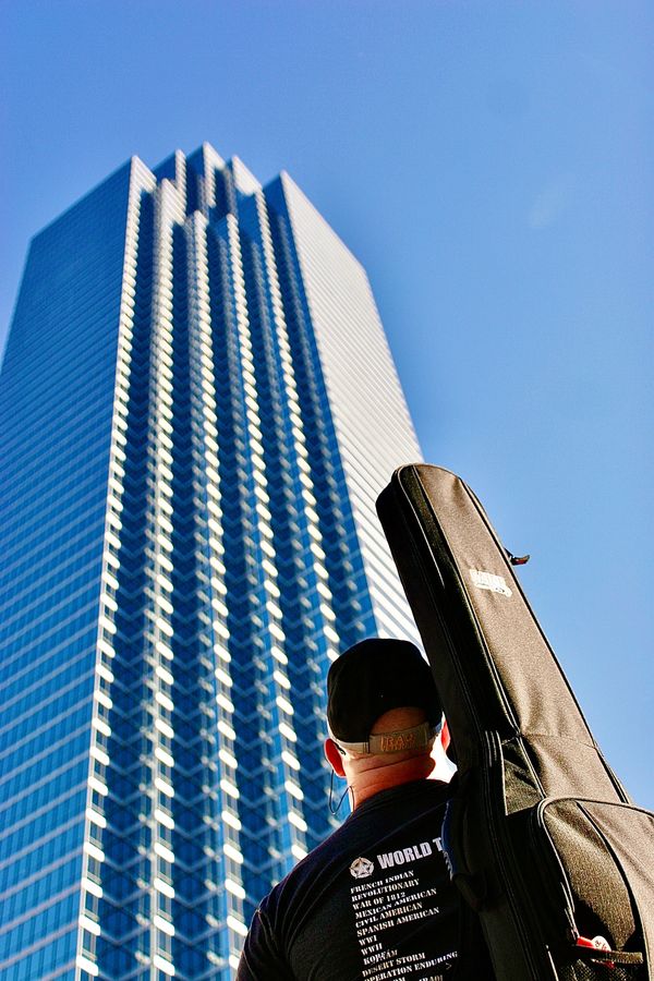 A guy in downtown Dallas, Tx with an electric guitar on his back looking up at a tall building