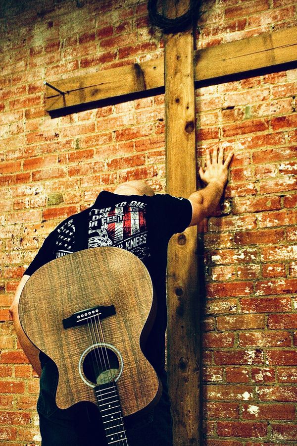 A guy with a guitar on his back and head bowed leaning against a brick wall in front of a cross