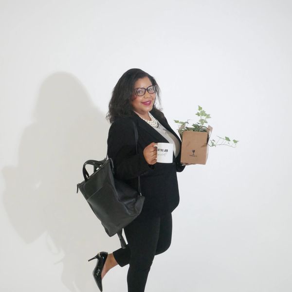 Woman in business attire holding a coffee mug and a plant, smiling confidently, walking into her first day on the job.