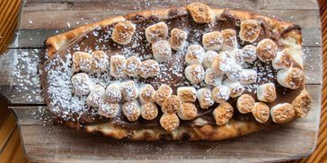 Flatbread topped with melted chocolate, toasted marshmallows, and powdered sugar on a wooden board.