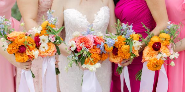 Bridal party holding vibrant orange and pink floral bouquets with white ribbons.