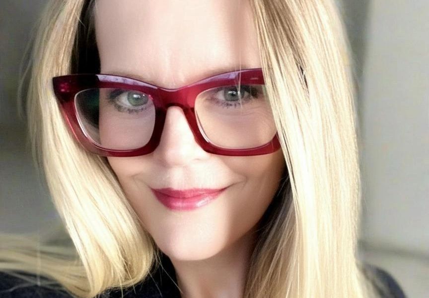 Blond woman with red glasses and red lipstick, smiling.