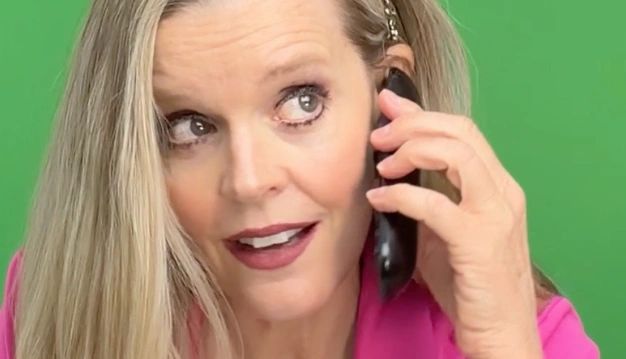 Blond woman in front of a green screen holding a black phone, wearing a pink blazer, smiling.