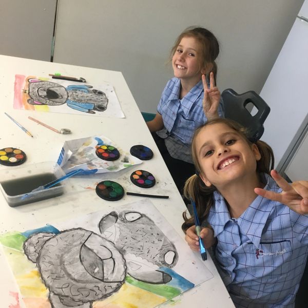 Two girls in blue school uniforms smiling and making peace signs while painting.