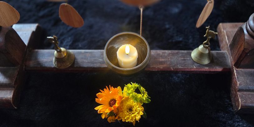 A lit candle and yellow flowers on a wooden platform with small brass bells.