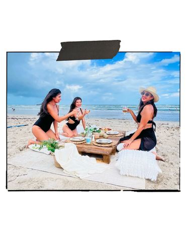 Three women enjoying a beach picnic with drinks and snacks under a blue sky.