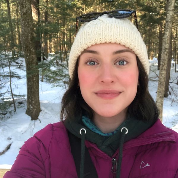A woman wearing a knit hat and jacket takes a selfie in a snowy forest.