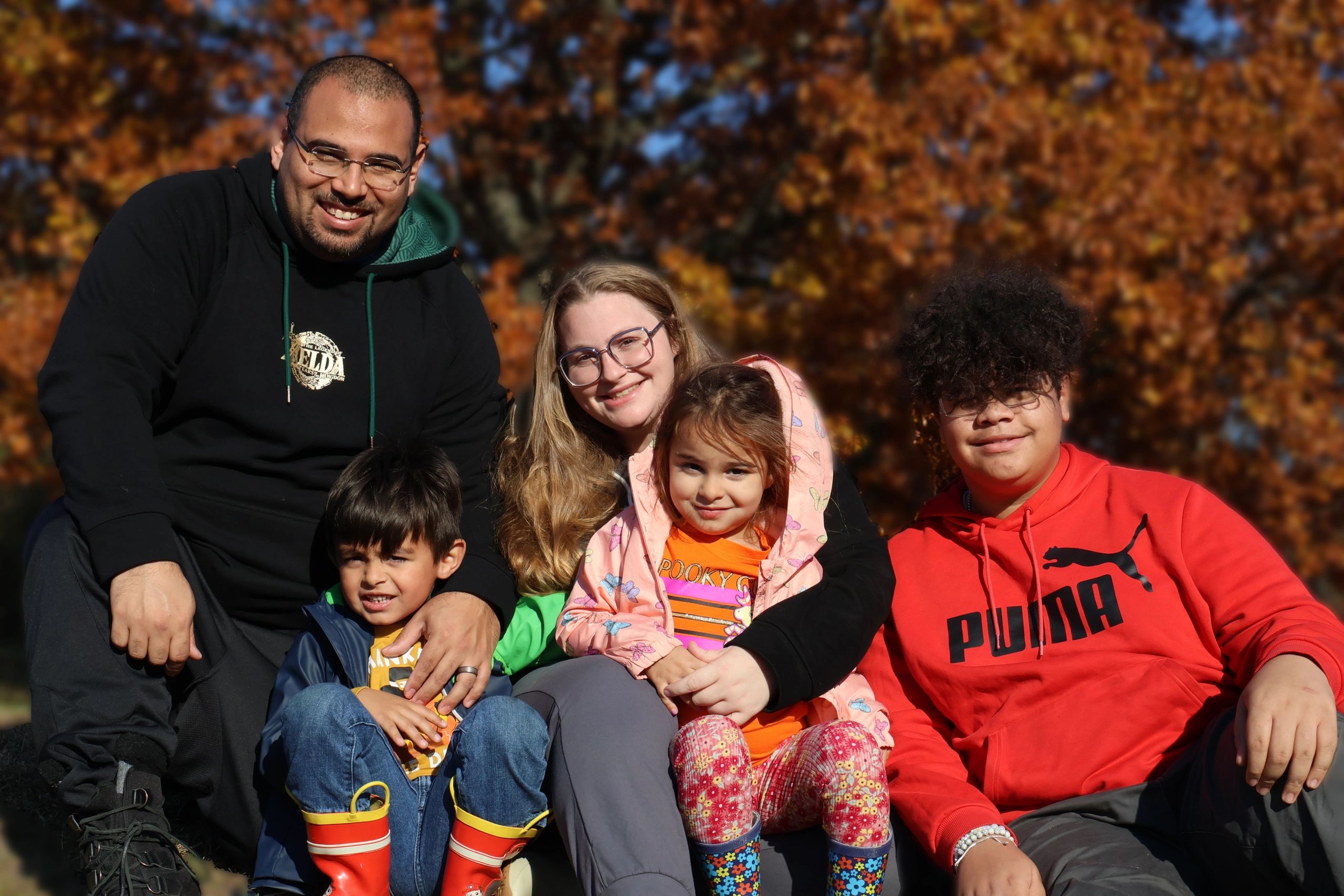 A smiling family poses outdoors with autumn leaves in the background.