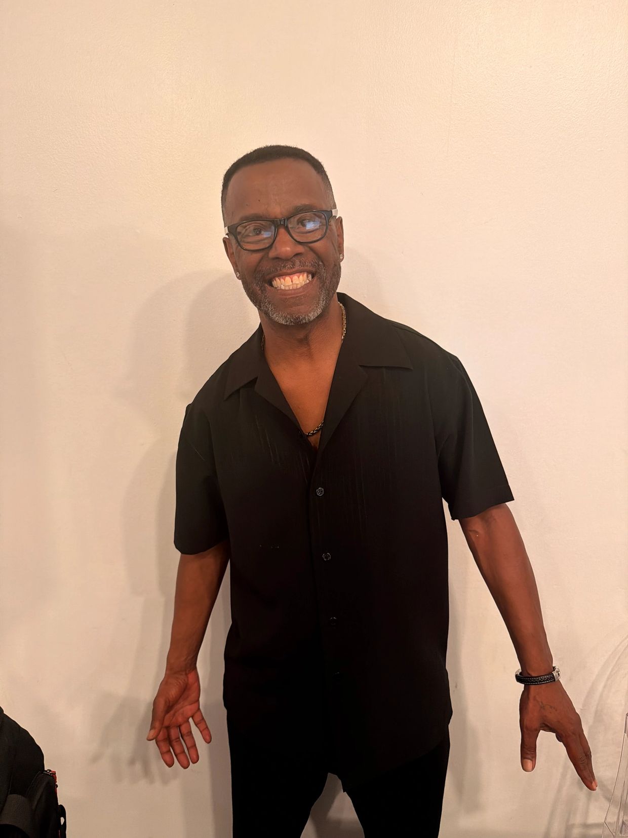A smiling man in a black shirt and glasses stands against a plain wall.