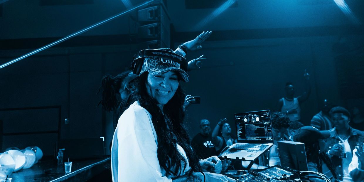 DJ wearing headphones and cap, smiling while mixing music at a lively club.