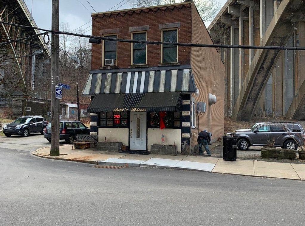 Small brick diner on a corner street under a bridge with parked cars nearby.