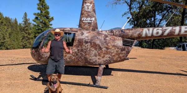 Man in sunglasses and hat with thumbs up by camo helicopter and dog.