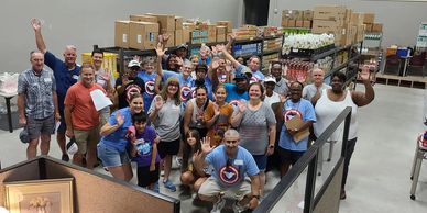 A diverse group of volunteers waving inside a warehouse filled with supplies.