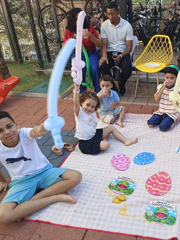 Children playing with balloon swords and sitting on a picnic blanket outdoors.