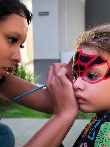 A woman paints a child's face with a Spider-Man design outdoors.