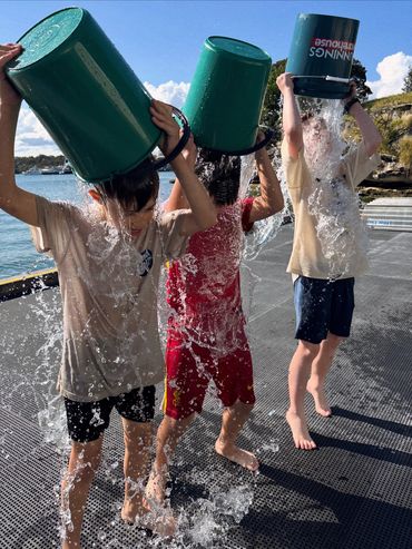Three kids dumping water on their heads with green buckets by the water.