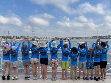 Group of children in matching blue shirts waving by the waterfront on a sunny day.