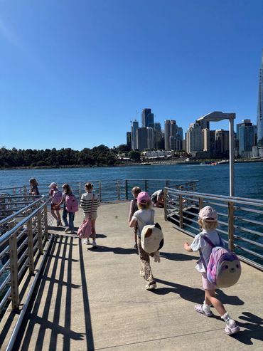 Children with colorful backpacks walk on a sunny pier by the water with a city skyline in the background.