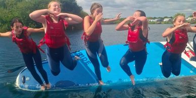Five girls in life jackets jump off a floating platform into the water.