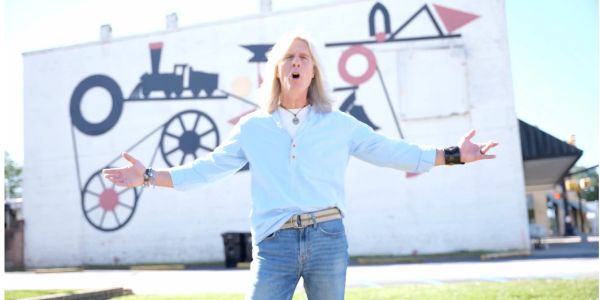 Man with blond hair singing outdoors in front of a mural.