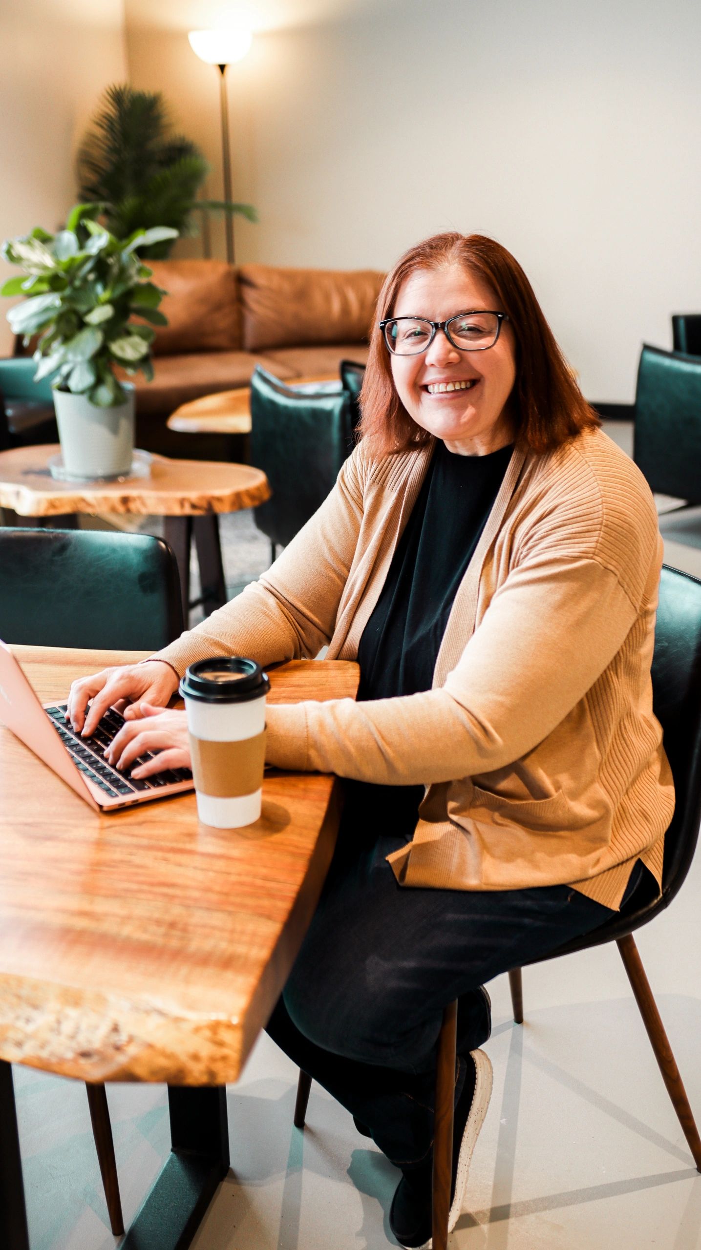 Picture of Dr. White at table typing on a laptop with a cup of coffee there as well. Couches and a p