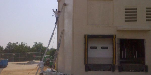 Two workers on a tall ladder beside a warehouse loading dock.