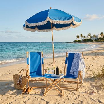 Two blue beach chairs and an umbrella set up on a sandy shore with ocean views.