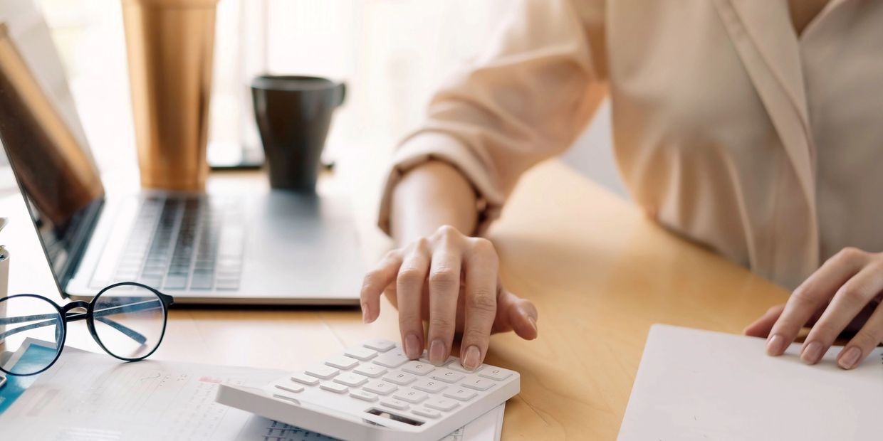 Person using a calculator at a desk with a laptop and glasses nearby.