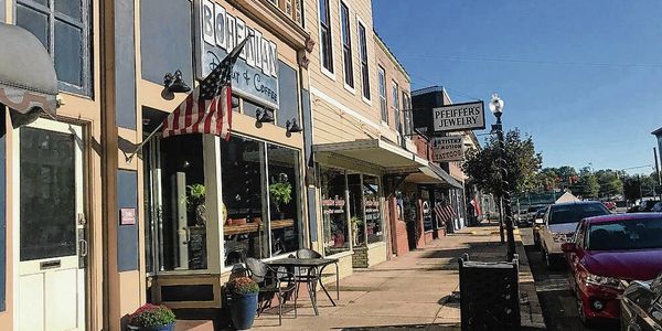 Quiet small-town street with shops, flags, and parked cars on a sunny day.