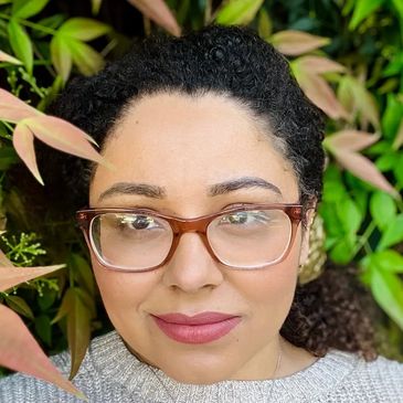 Woman with glasses smiling outdoors surrounded by green leaves.