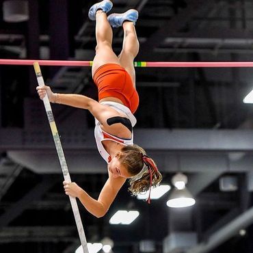 A female athlete in mid-air during a pole vault, wearing a red and white uniform