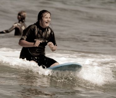 A women wearing a wetsuit joyfully surfs on a wave with a light blue surfboard.
