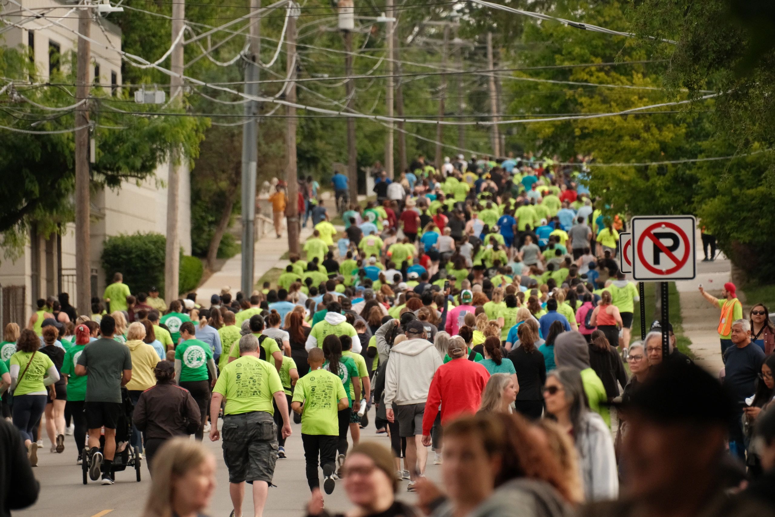 Large crowd participating in an outdoor walk or run event along a tree-lined street.