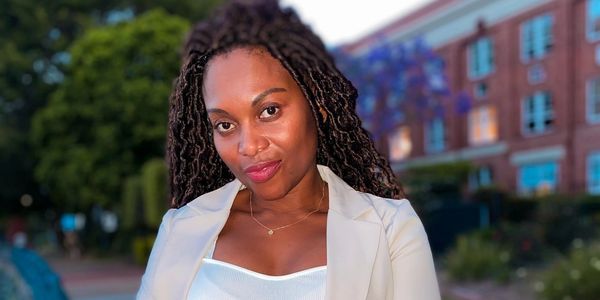 A confident woman with braided hair smiles outdoors in evening light.