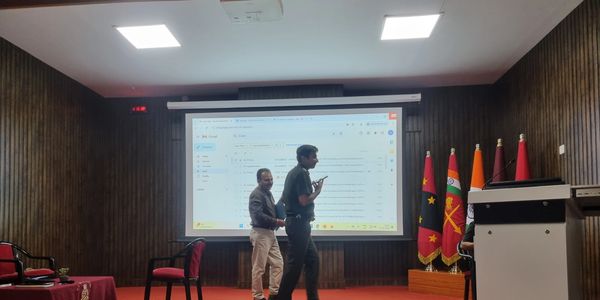 Two men on stage with a projection screen and flags in a conference room.
