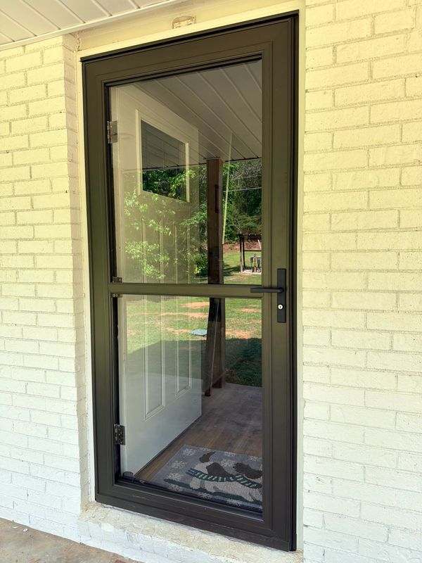 A black-framed glass storm door on a white brick wall with a white door behind it.